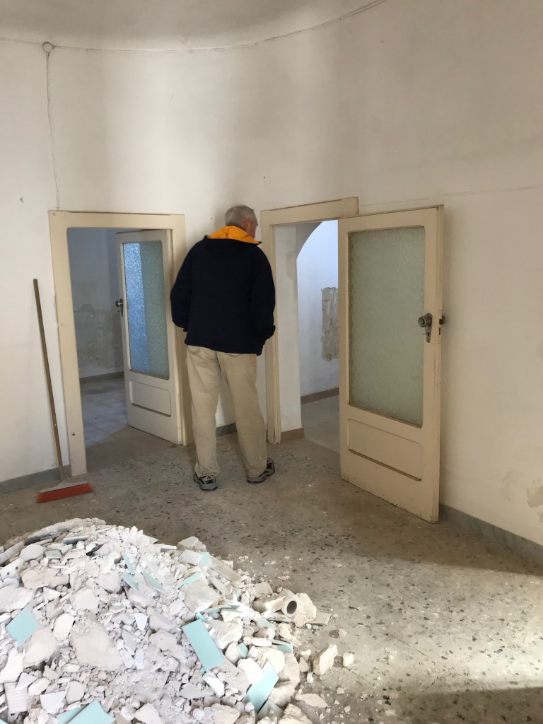 Man looking through door of trullo with pile of trash behind him